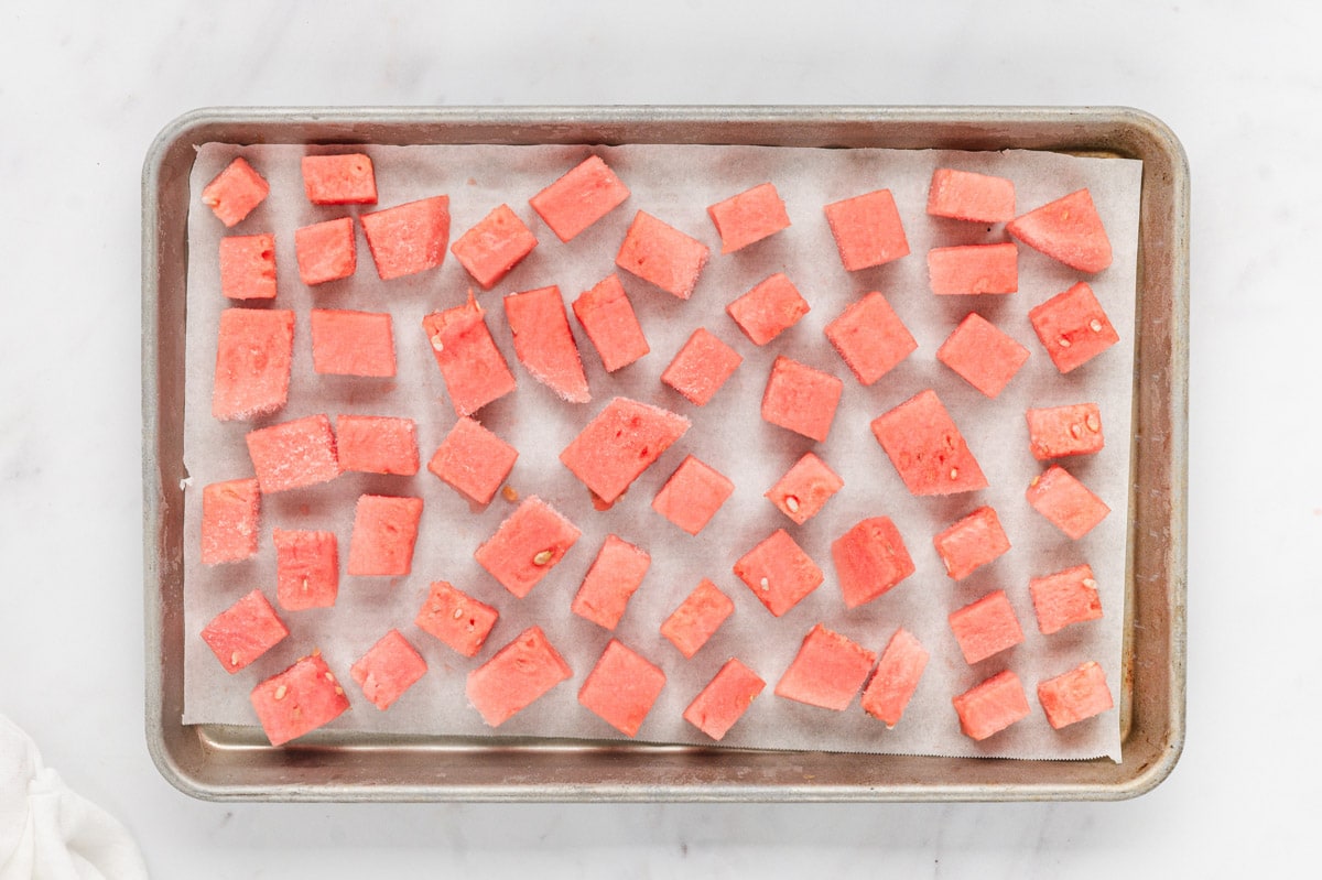 Evenly cut watermelon cubes spread in a single layer on a parchment-lined baking sheet on a kitchen counter