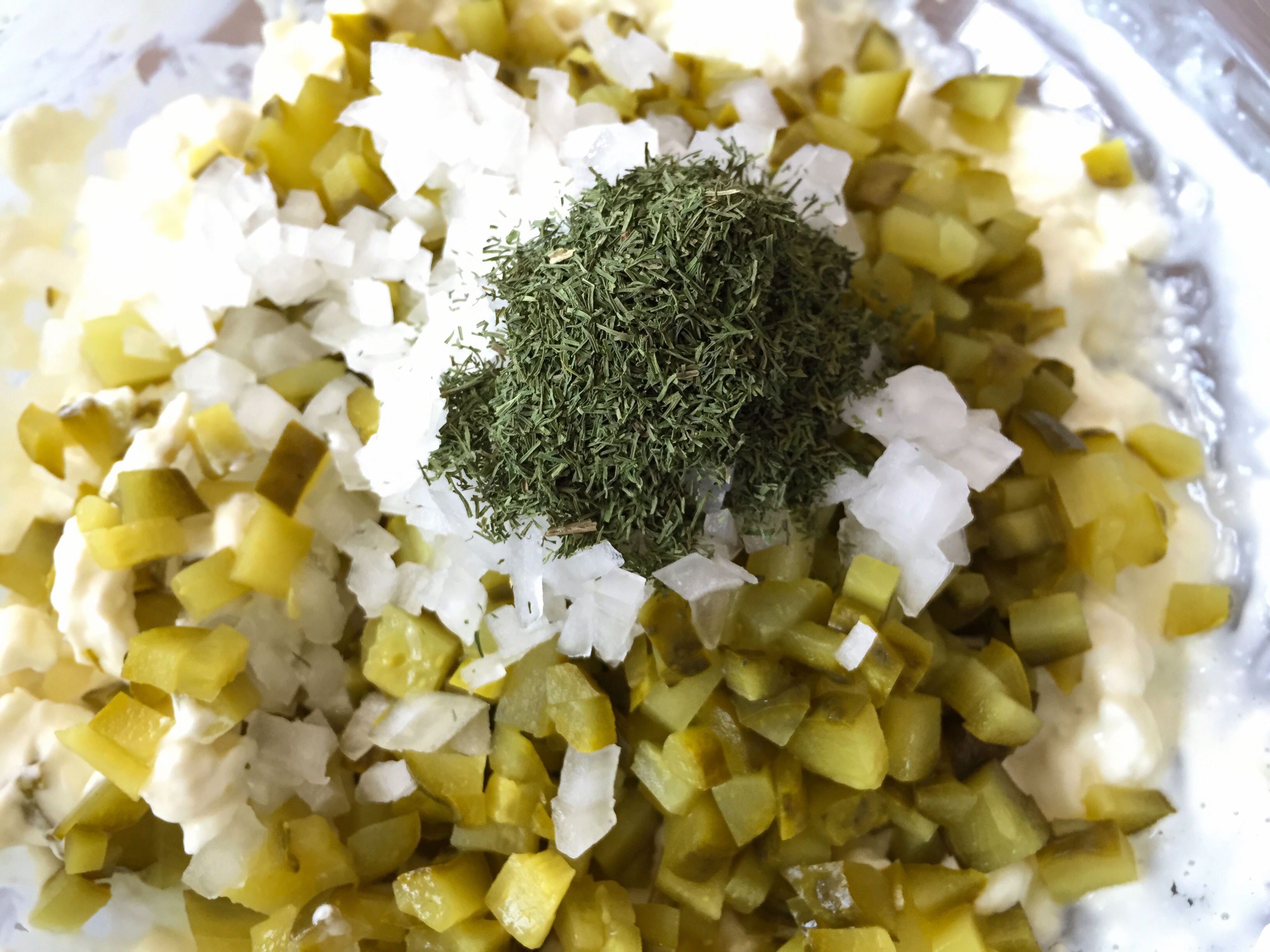 Finely chopped dill pickles and minced onion on a cutting board beside measuring spoons on a kitchen counter