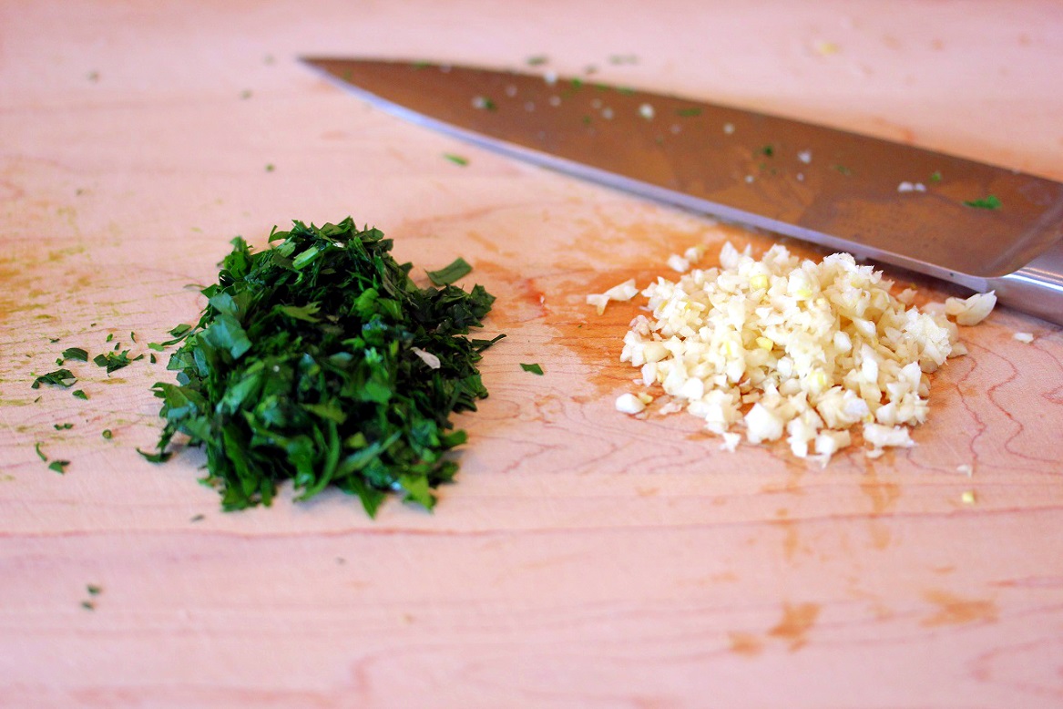 Finely chopped flat-leaf parsley and minced garlic on a wooden cutting board with a chef’s knife beside them