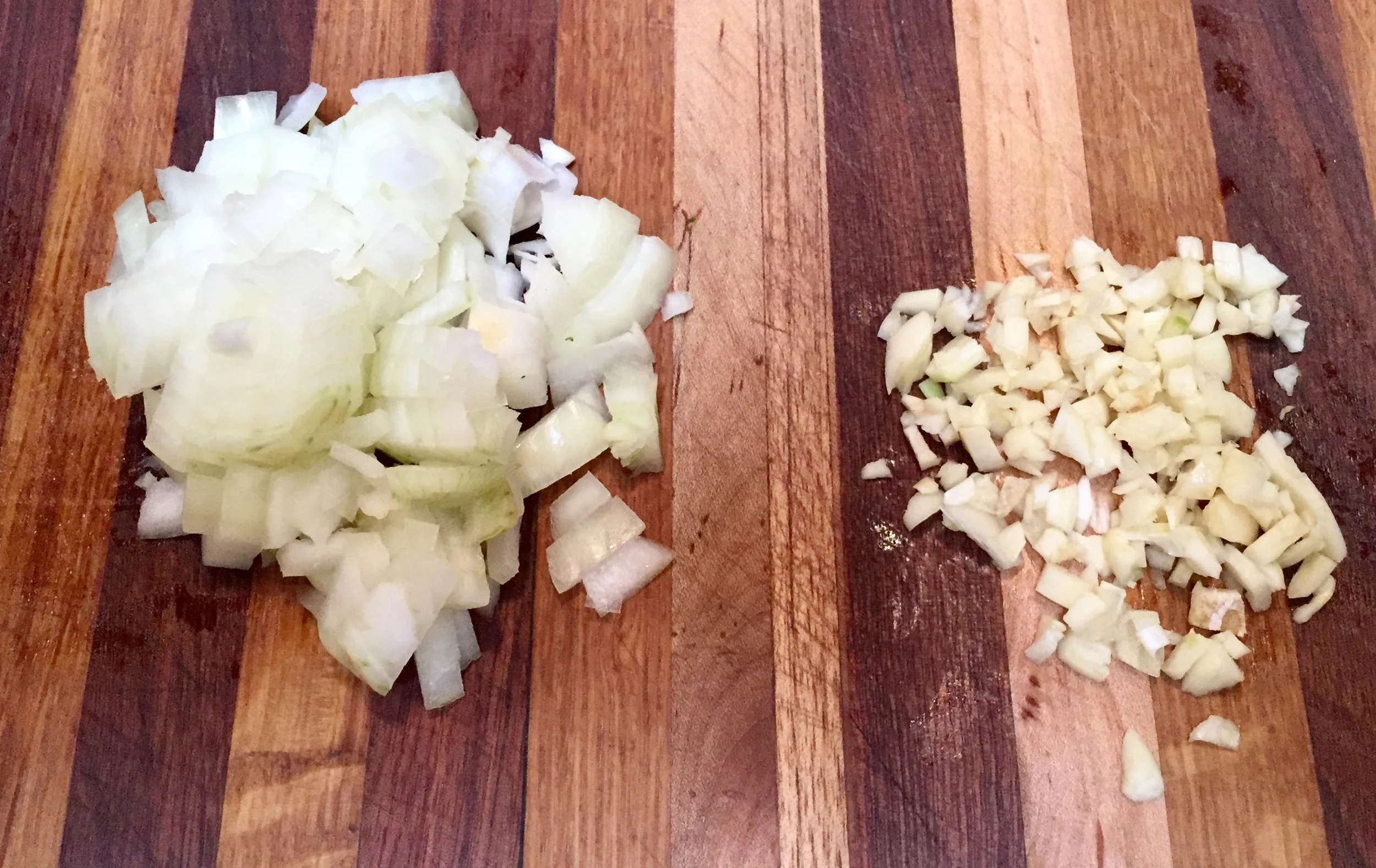 Finely chopped onion and minced garlic on a wooden cutting board with a chef's knife, set on a clean kitchen counter