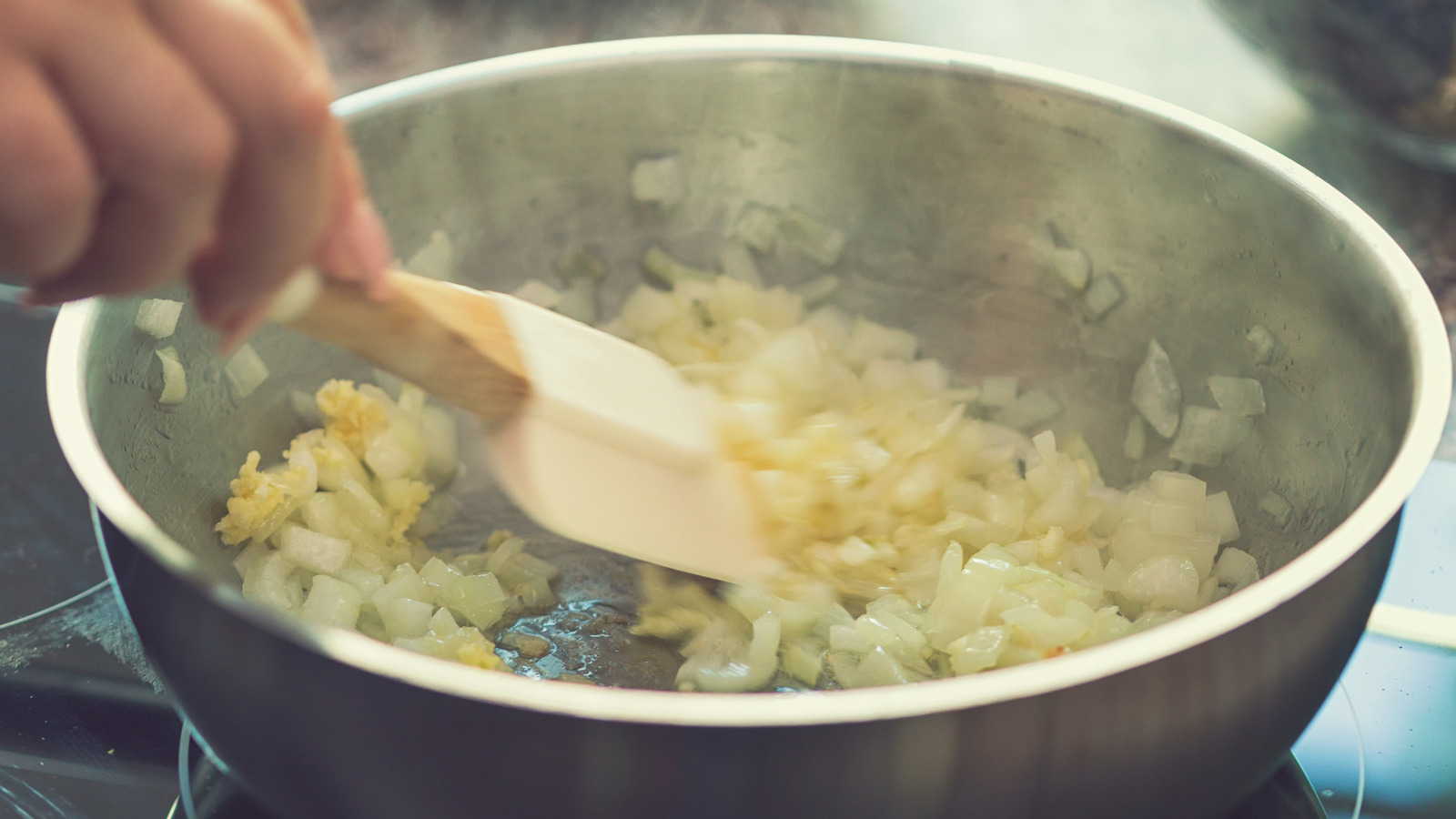 Finely chopped onions and minced garlic softening in a saucepan on the stovetop while being stirred with a wooden spoon