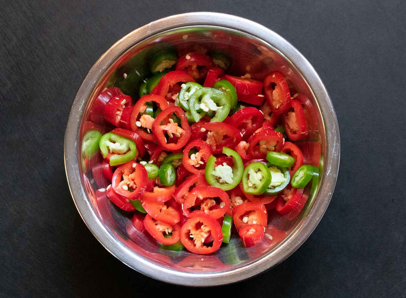 Finely chopped red and green peppers and jalapeños on a wooden cutting board next to a chef's knife