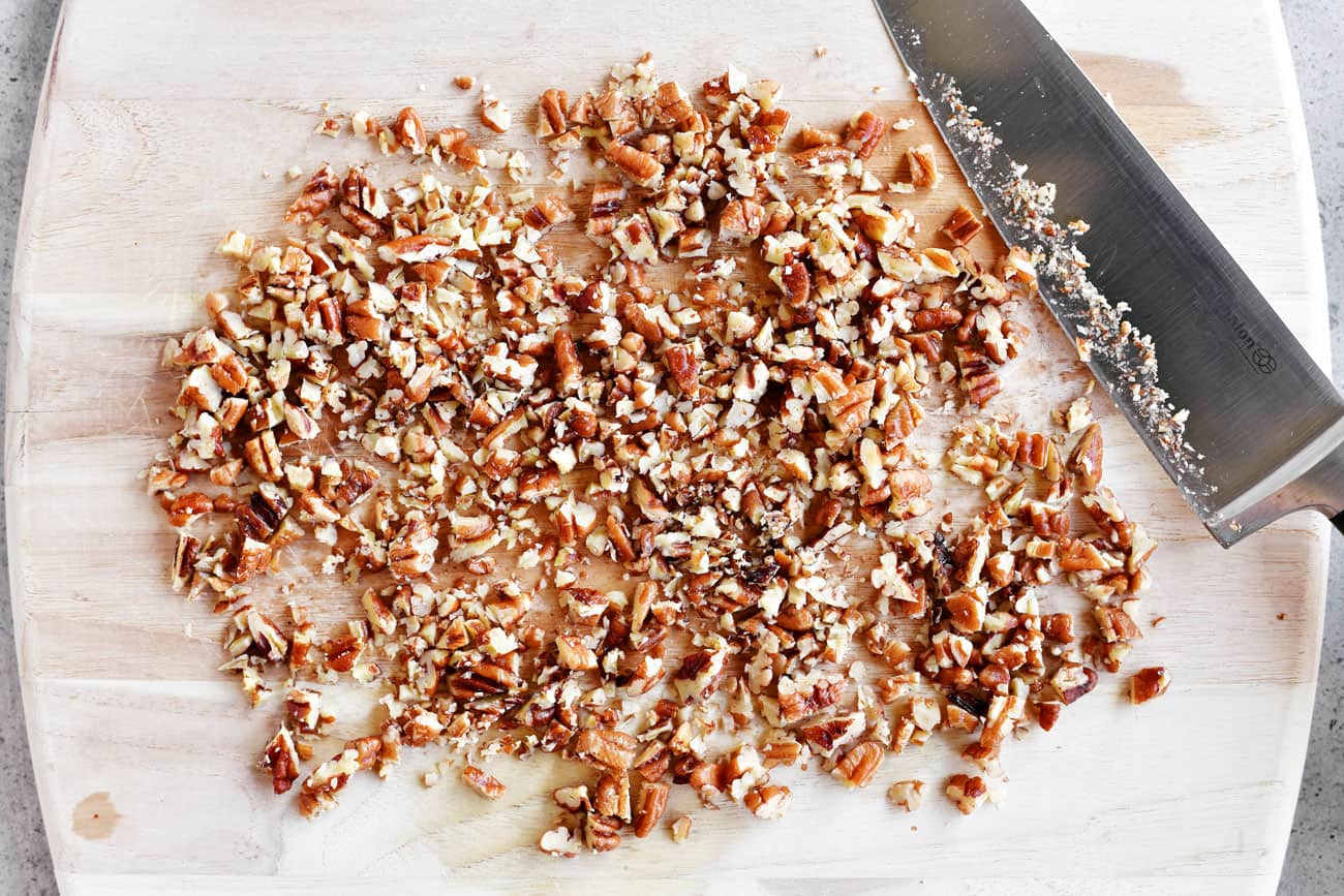 Finely chopped toasted pecans piled on a wooden cutting board beside a chef's knife on a kitchen counter