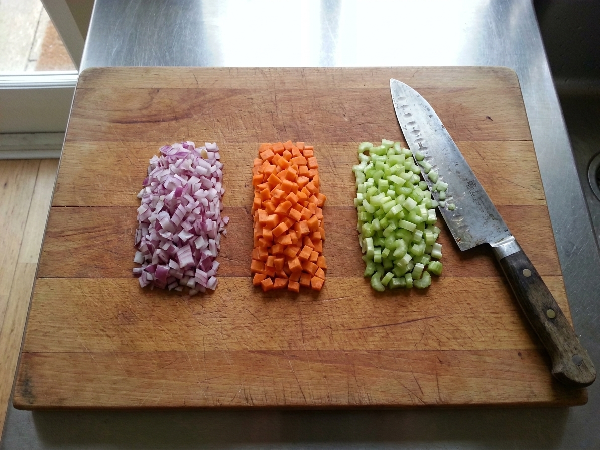 Finely diced onion, carrot, and celery in small piles on a wooden cutting board with a chef’s knife beside them