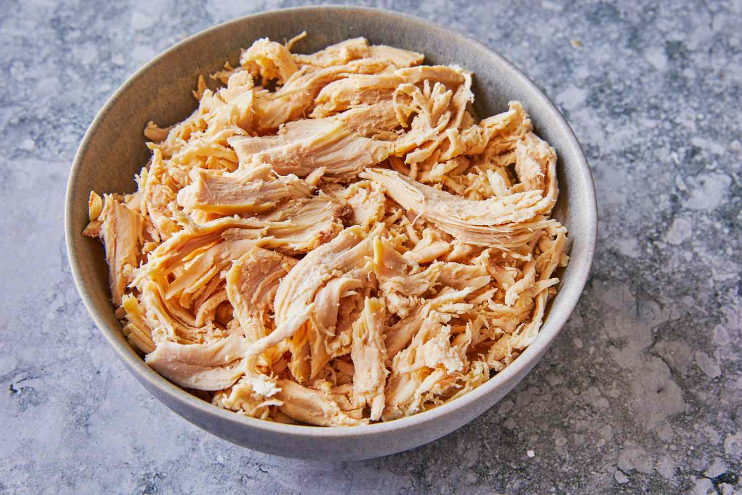Finely shredded cooked chicken in a large mixing bowl on a kitchen counter next to a wooden spoon