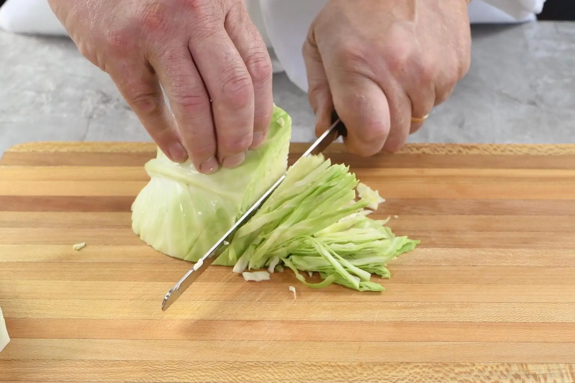 Finely shredded green cabbage piled on a wooden cutting board on a bright kitchen counter, with a chef's knife nearby