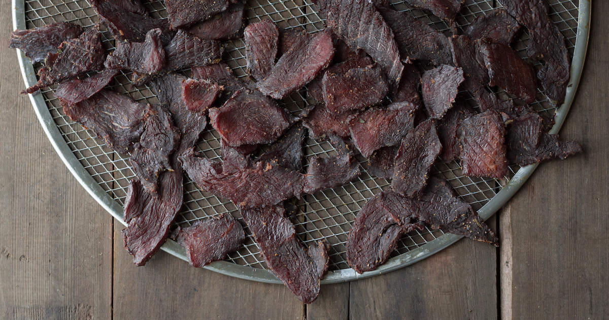 Finished deer jerky strips drying on a wire rack over a sheet pan on a neutral stone kitchen counter, warm natural window light