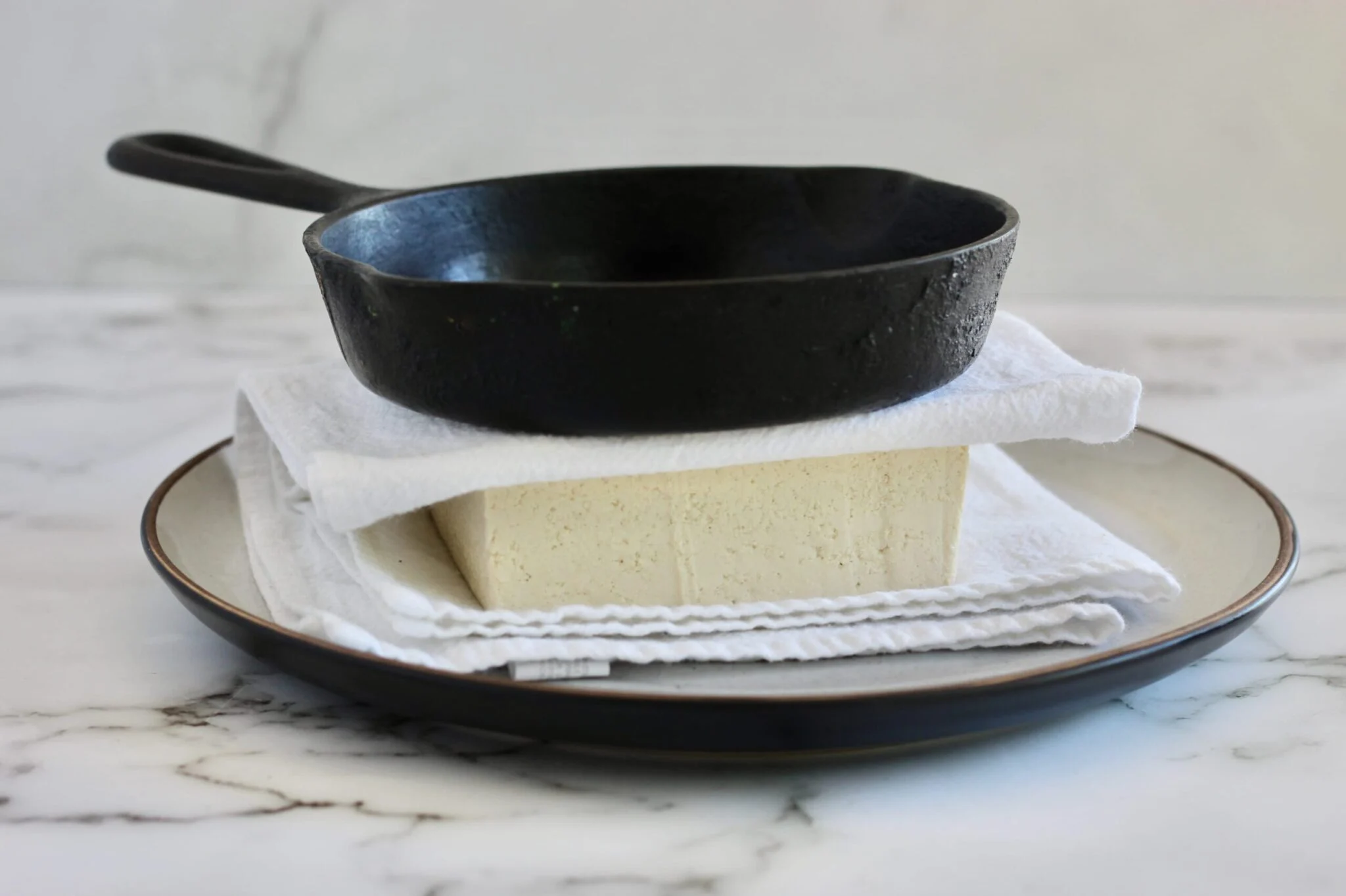 Firm tofu wrapped in a kitchen towel with a cast iron skillet set on top to press it on a plate on a kitchen counter
