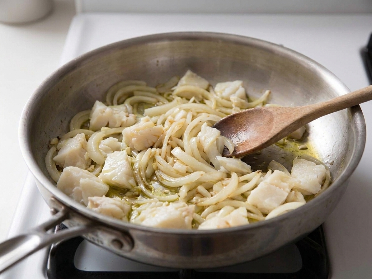 Flaked salt cod being stirred into sautéed onions and garlic in a skillet