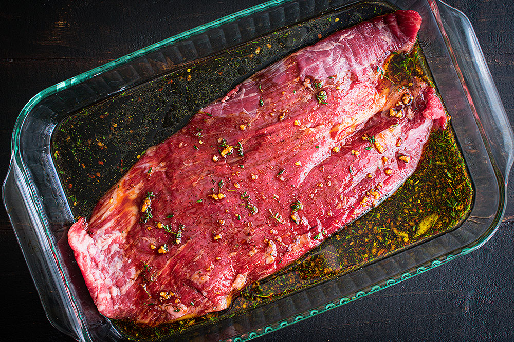 Flank steak in a glass dish being coated with a citrus-garlic marinade on a kitchen counter