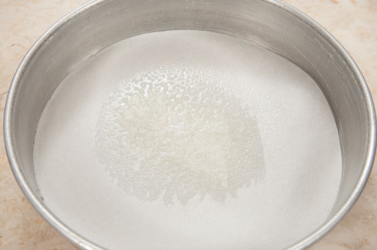 Flour being measured into a bowl beside two parchment-lined 8-inch cake pans on a clean kitchen counter
