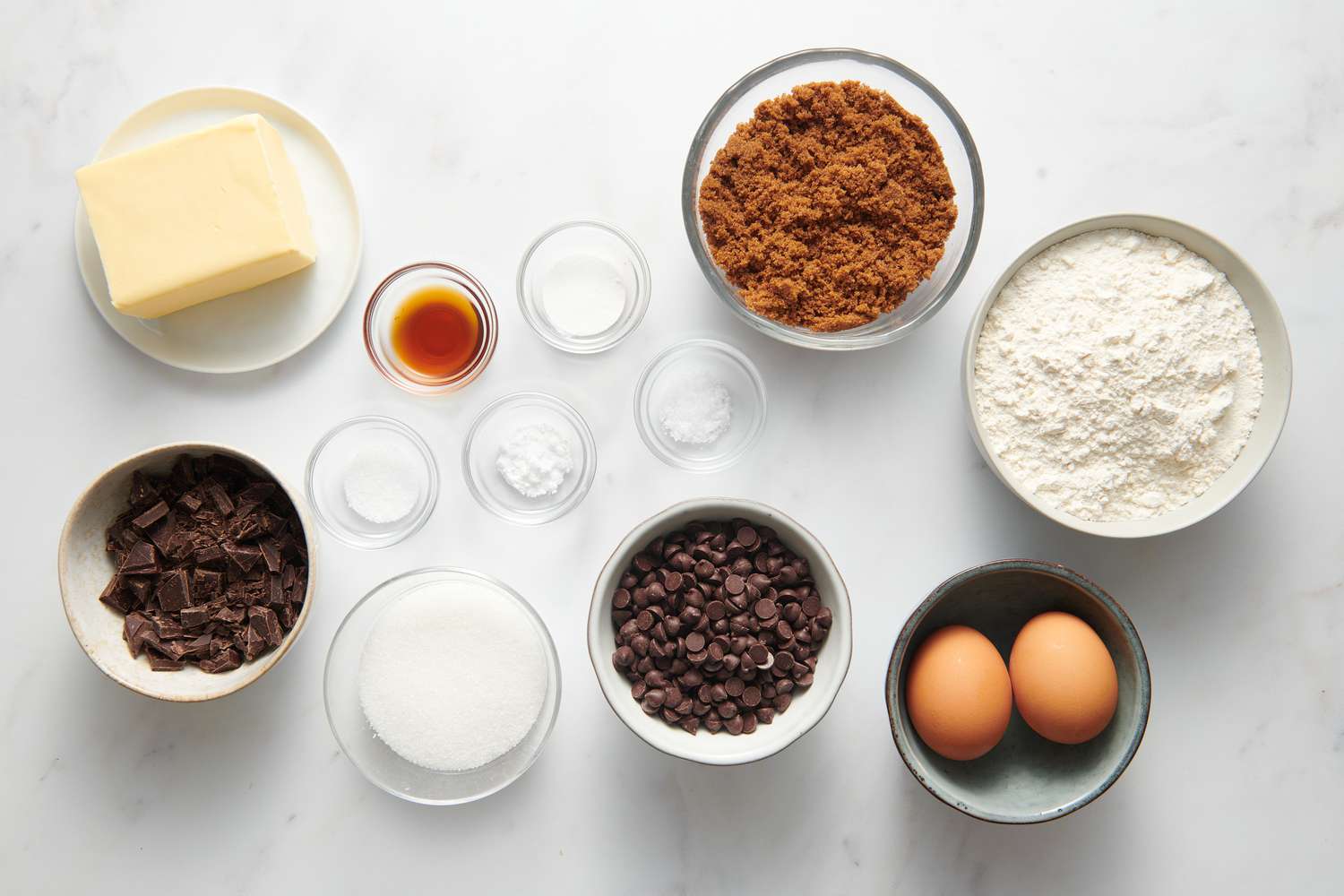 Flour, brown sugar, white sugar, butter, eggs, vanilla, baking soda, salt, and Ghirardelli chocolate chips arranged on a bright kitchen counter in small bowls