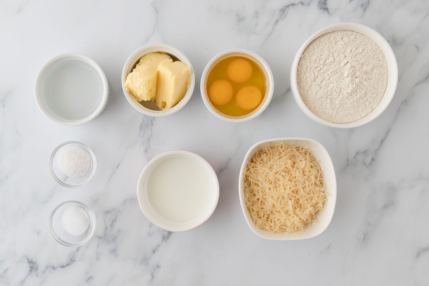 Flour, butter, eggs, milk, water, salt, and sugar measured in small bowls on a clean kitchen counter, ready for choux pastry