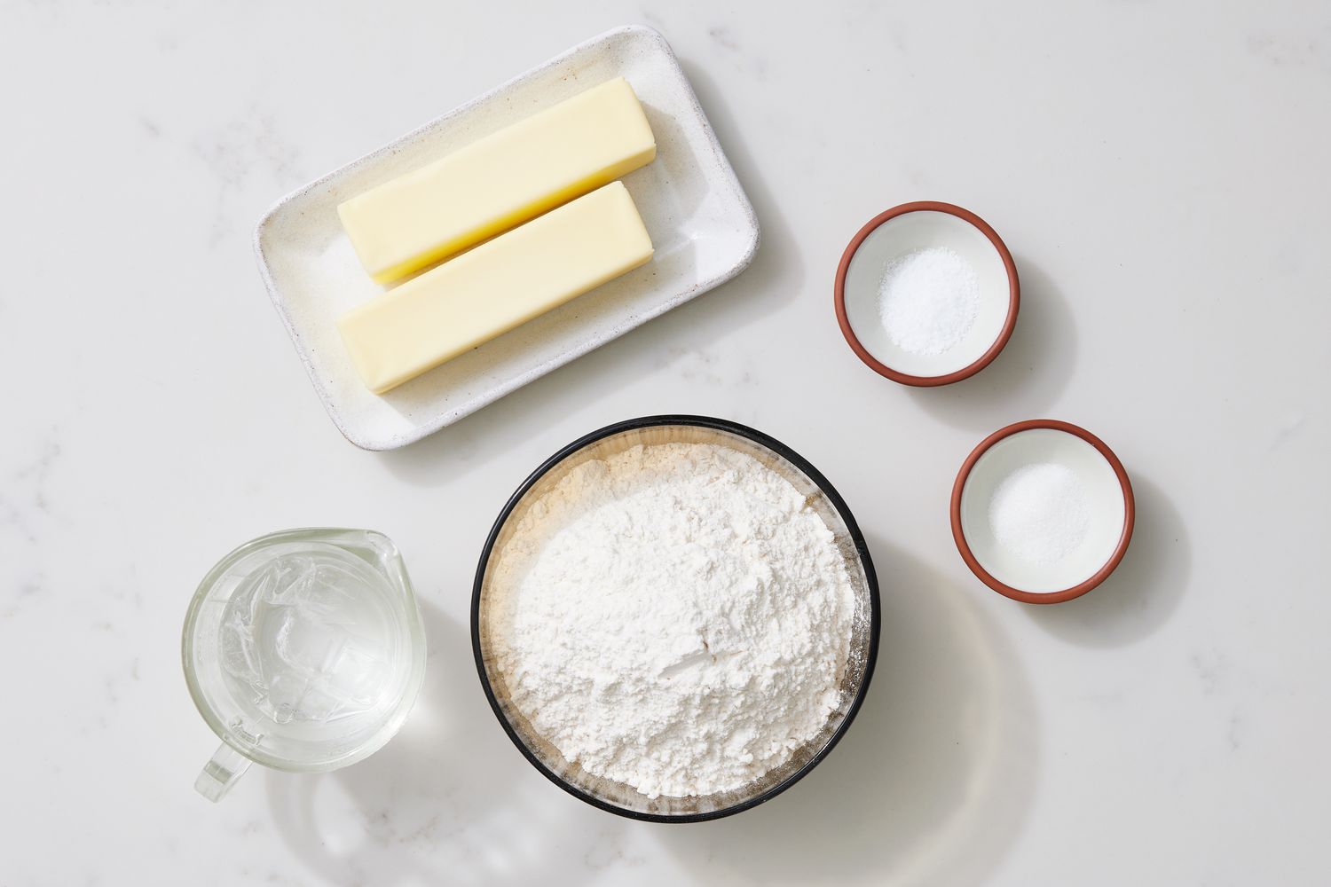 Flour, cold butter cubes, salt, and a small bowl of ice water arranged on a kitchen counter for pie dough