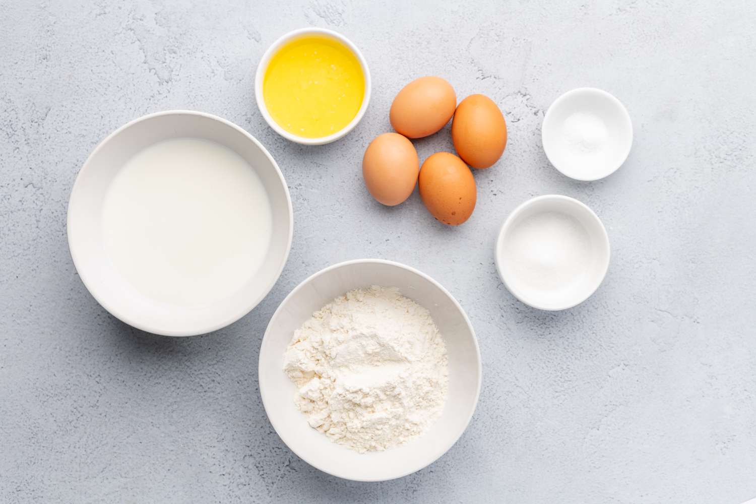 Flour, eggs, milk, water, melted butter, salt, and sugar arranged on a bright kitchen counter with measuring cups