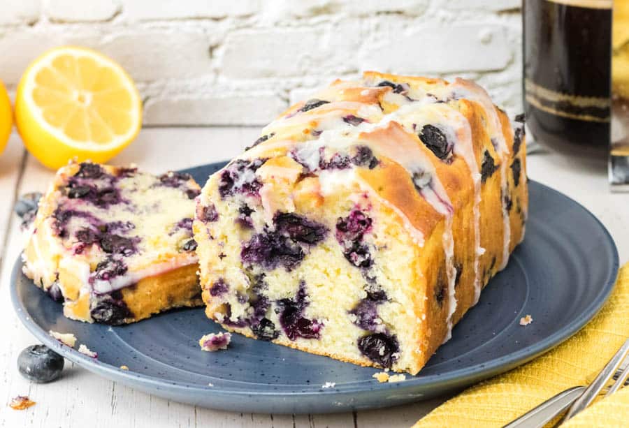 Flour, sugar, lemons, blueberries, eggs, butter, milk, and a loaf pan arranged on a clean kitchen counter in natural light