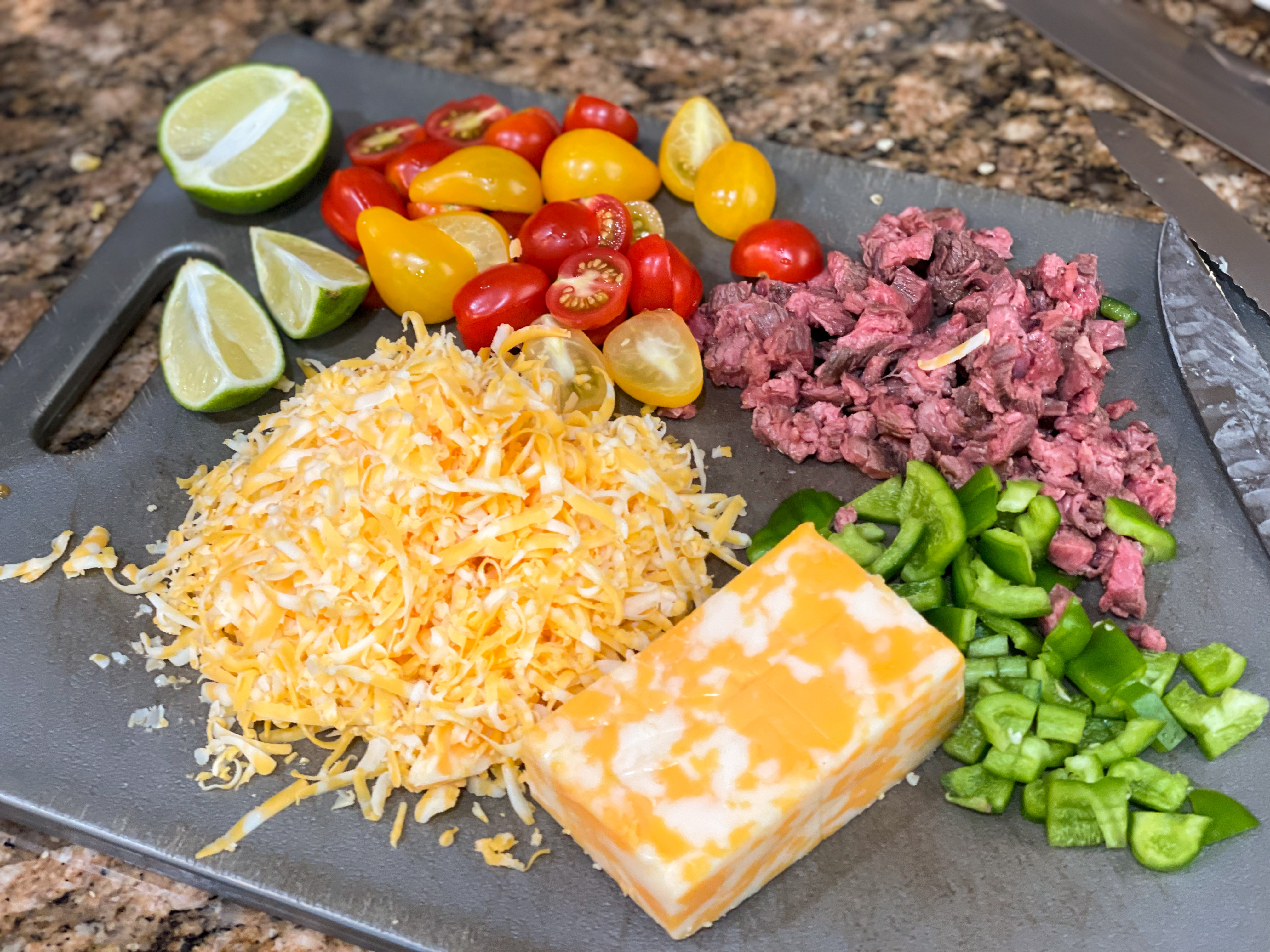 Flour tortillas, shredded cheese, sliced steak, bell peppers, onion, lime, and small bowls of spices arranged on a wooden cutting board on a bright kitchen counter