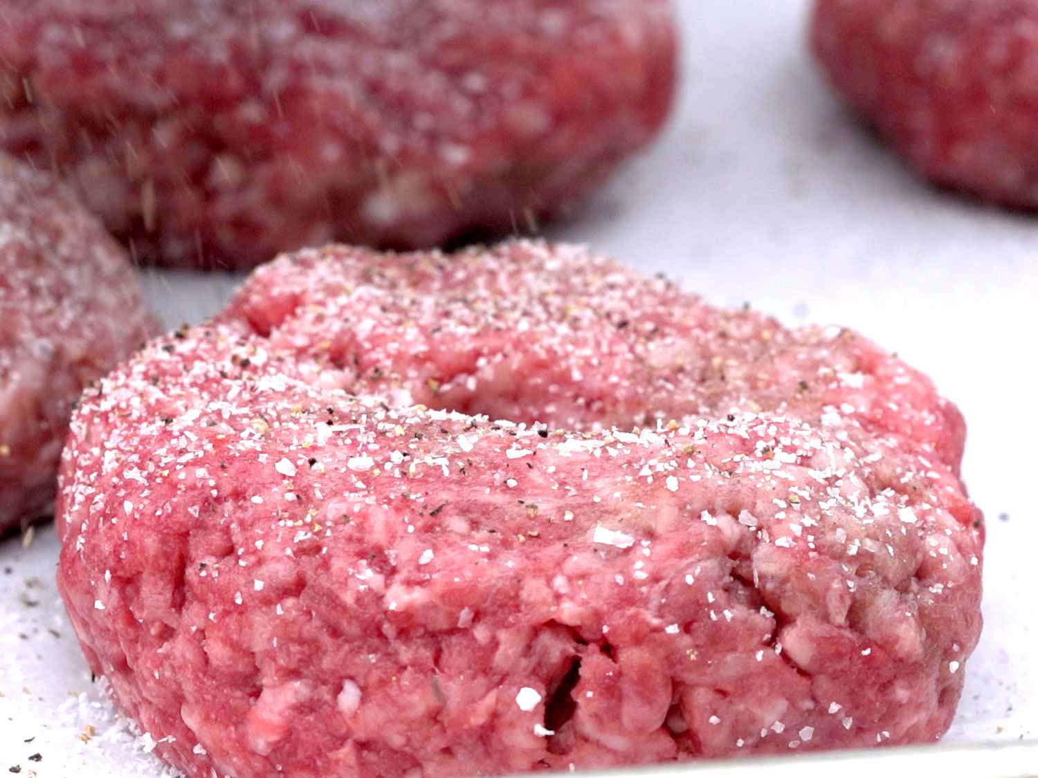 Four raw hamburger patties on parchment paper on a baking sheet, each with a small indentation in the center, under warm kitchen lighting