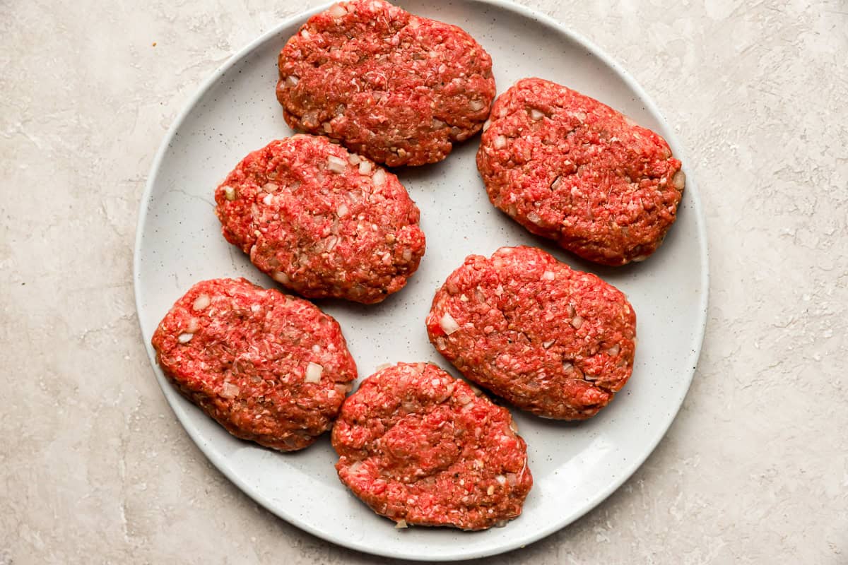 Four raw oval-shaped Hamburg steak patties with small center dimples resting on a plate beside sliced onions