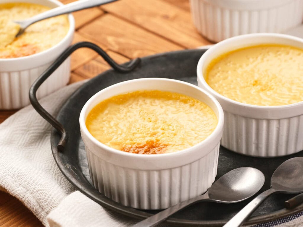 Four small ramekins of baked egg custard with lightly browned tops and a dusting of nutmeg on a neutral linen napkin on a warm kitchen counter, natural window light
