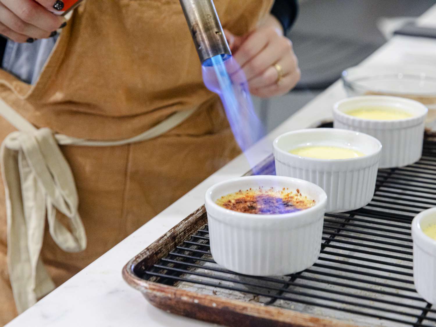 Four vanilla creme brulees in white ramekins with glassy caramelized sugar tops on a warm-toned kitchen counter, soft natural light