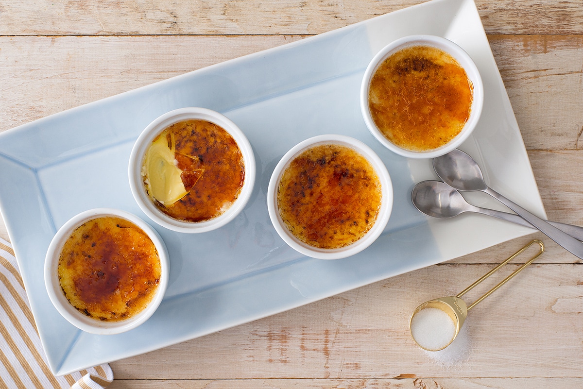 Four vanilla crème brûlées in white ramekins with glossy caramelized sugar tops on a light stone countertop, soft natural window light