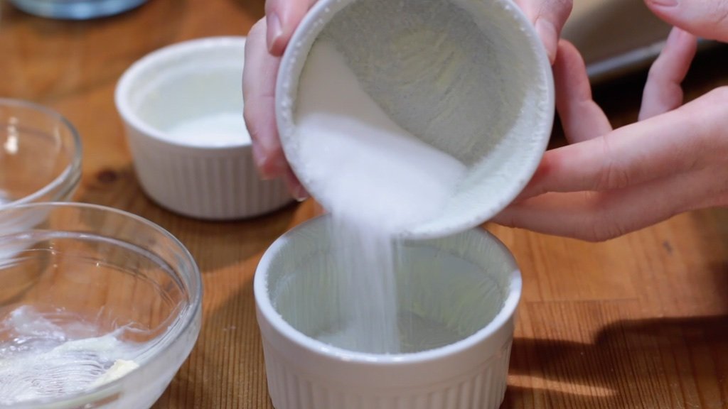 Four white ramekins coated with butter and a sugar lining, sitting on a baking sheet on a kitchen counter