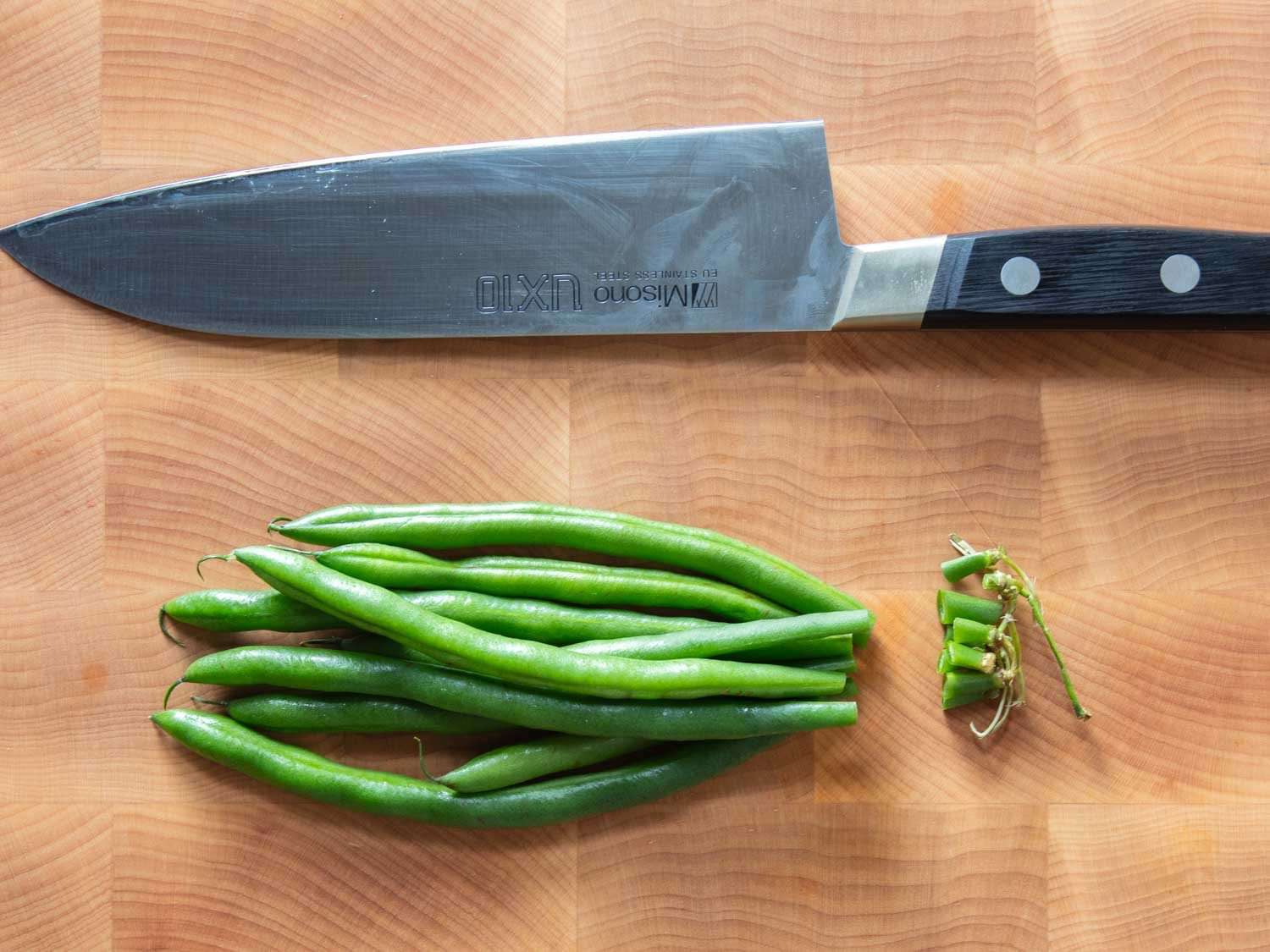 Fresh French green beans trimmed on a wooden cutting board with a chef’s knife and a small pile of cut stem ends nearby