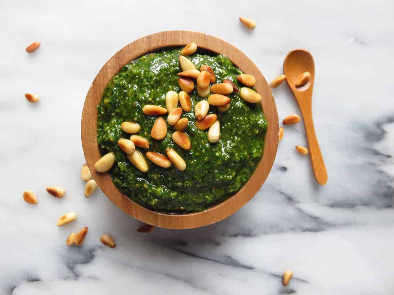 Fresh basil leaves, pine nuts, garlic cloves, grated Parmesan, and a lemon on a wooden cutting board in a home kitchen