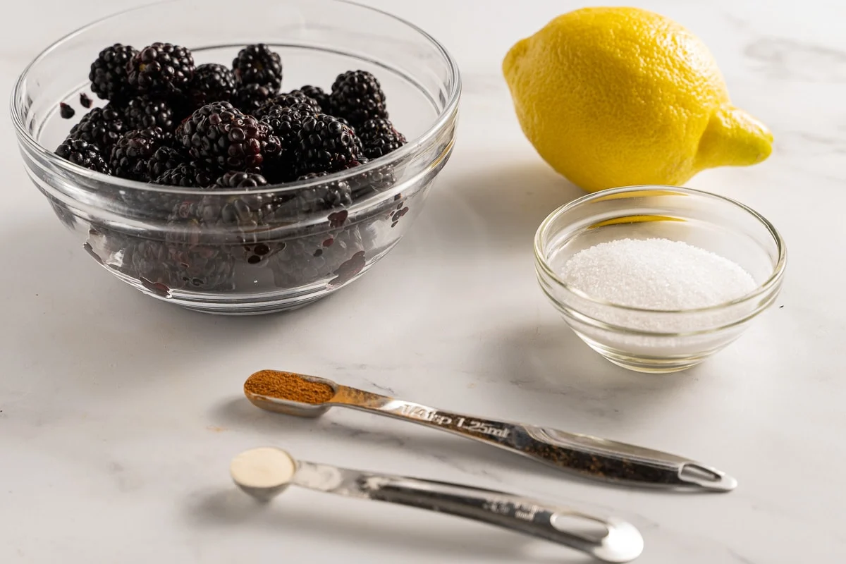 Fresh blackberries, a whole lemon, and a bowl of granulated sugar arranged on a clean kitchen counter, ready for jam making