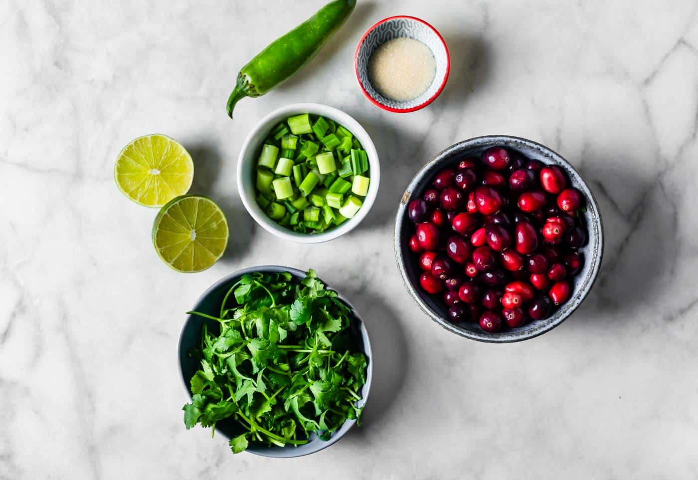 Fresh cranberries, a jalapeño, cilantro, green onions, limes, and sugar arranged on a clean kitchen counter in natural light