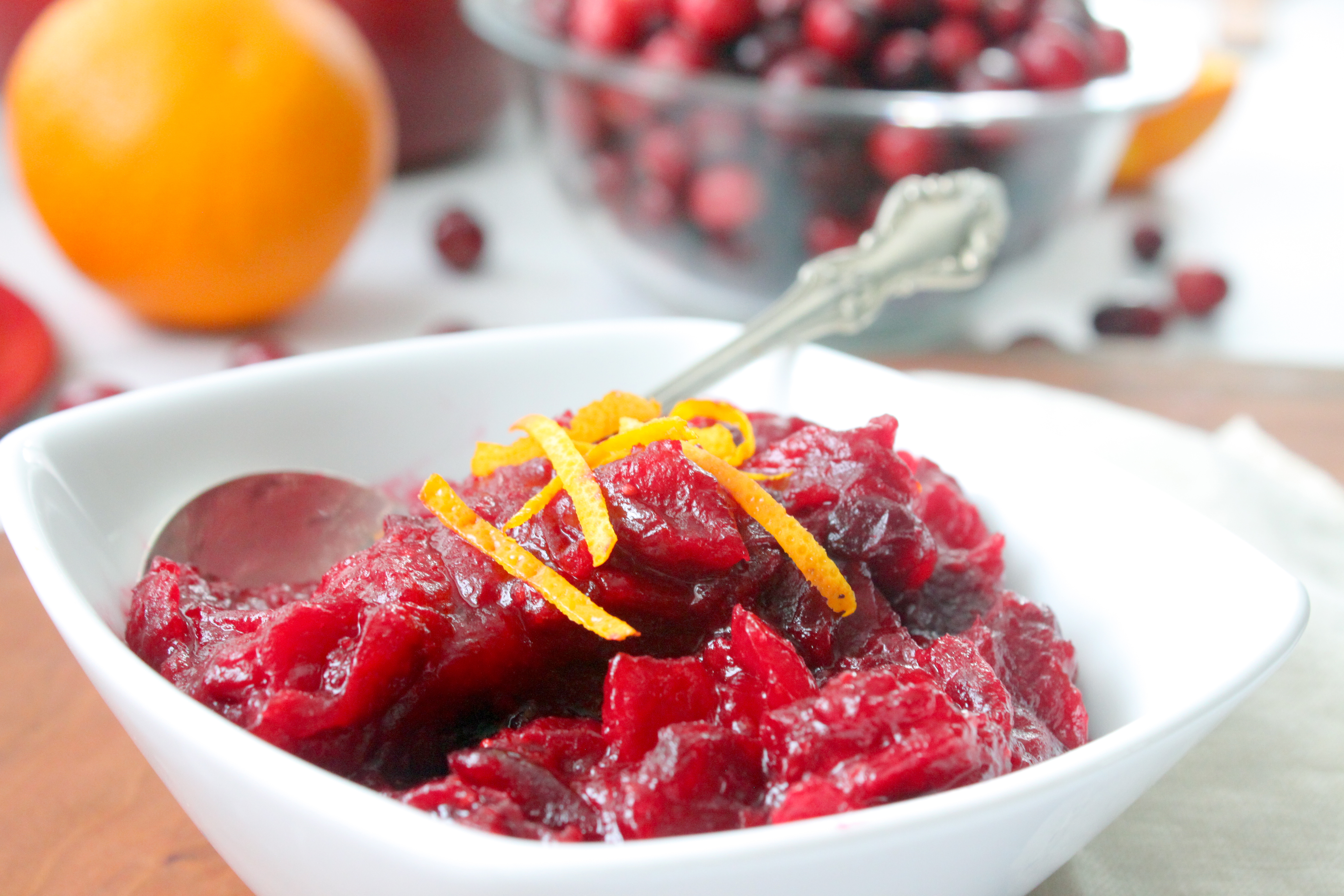 Fresh cranberries, a whole orange, and a small bowl of sugar arranged on a light kitchen counter, ready for cooking