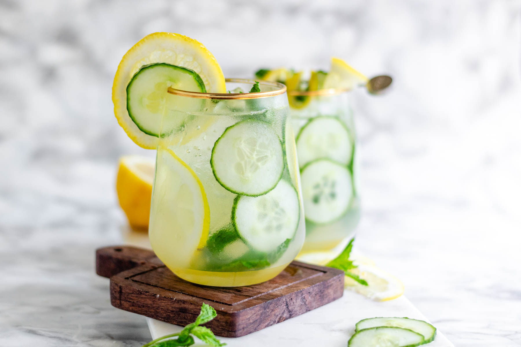 Fresh cucumber slices, mint sprigs, and lemon and orange slices prepared on a wooden cutting board beside a glass filled with ice