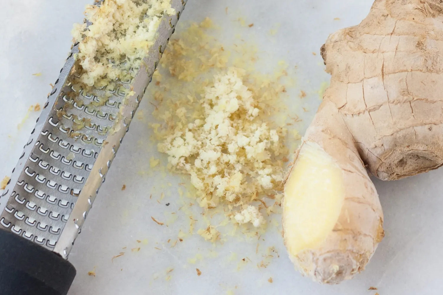 Fresh ginger being grated on a microplane into a small bowl on a clean kitchen counter