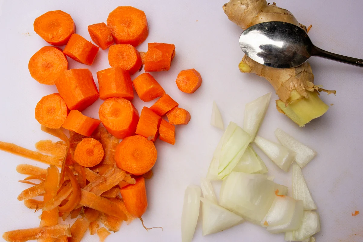 Fresh ginger root, a halved yellow onion, and peeled carrot pieces arranged on a wooden cutting board on a bright kitchen counter