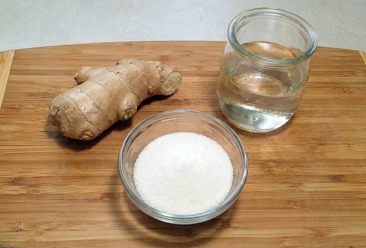 Fresh ginger root, a small bowl of sugar, and a jar of water on a clean kitchen counter