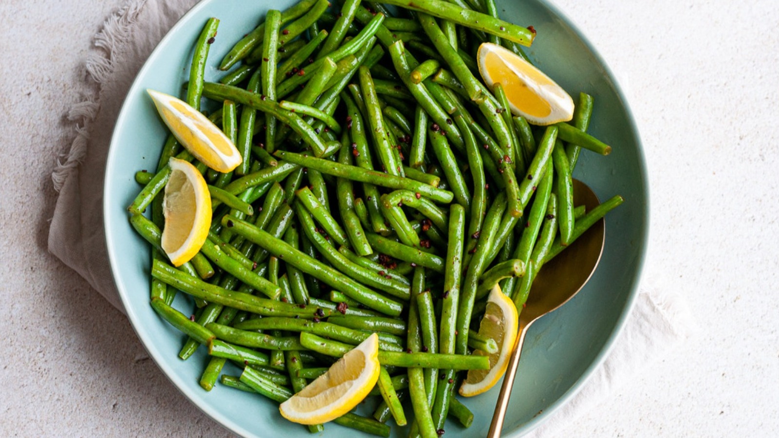 Fresh green beans, a whole lemon, garlic cloves, olive oil, salt, and pepper arranged on a cutting board on a kitchen counter