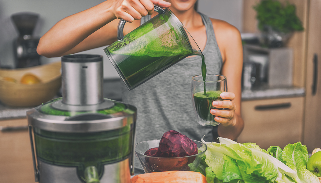 Fresh green juice pouring from a juicer spout into a clear glass measuring jug on a kitchen counter