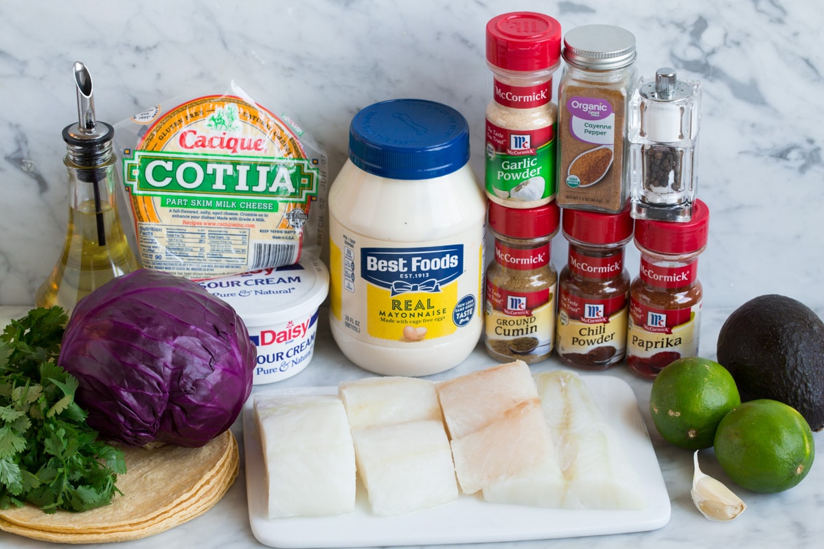 Fresh ingredients for fish tacos and coleslaw arranged on a bright kitchen counter including tortillas, cabbage, limes, spices, and white fish