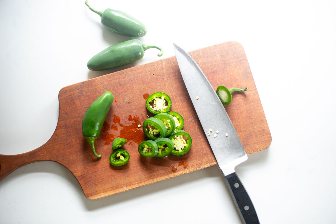 Fresh jalapeño being sliced into thin rounds on a wooden cutting board with a chef's knife
