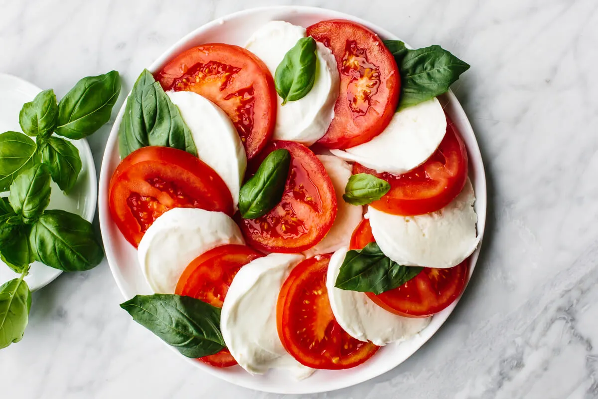Fresh mozzarella slices, cherry tomatoes, and basil leaves on a cutting board with a chef’s knife
