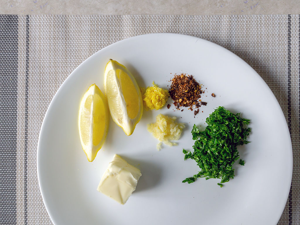 Fresh parsley, whole lemon, garlic cloves, flaky salt, and a microplane laid out on a light kitchen counter