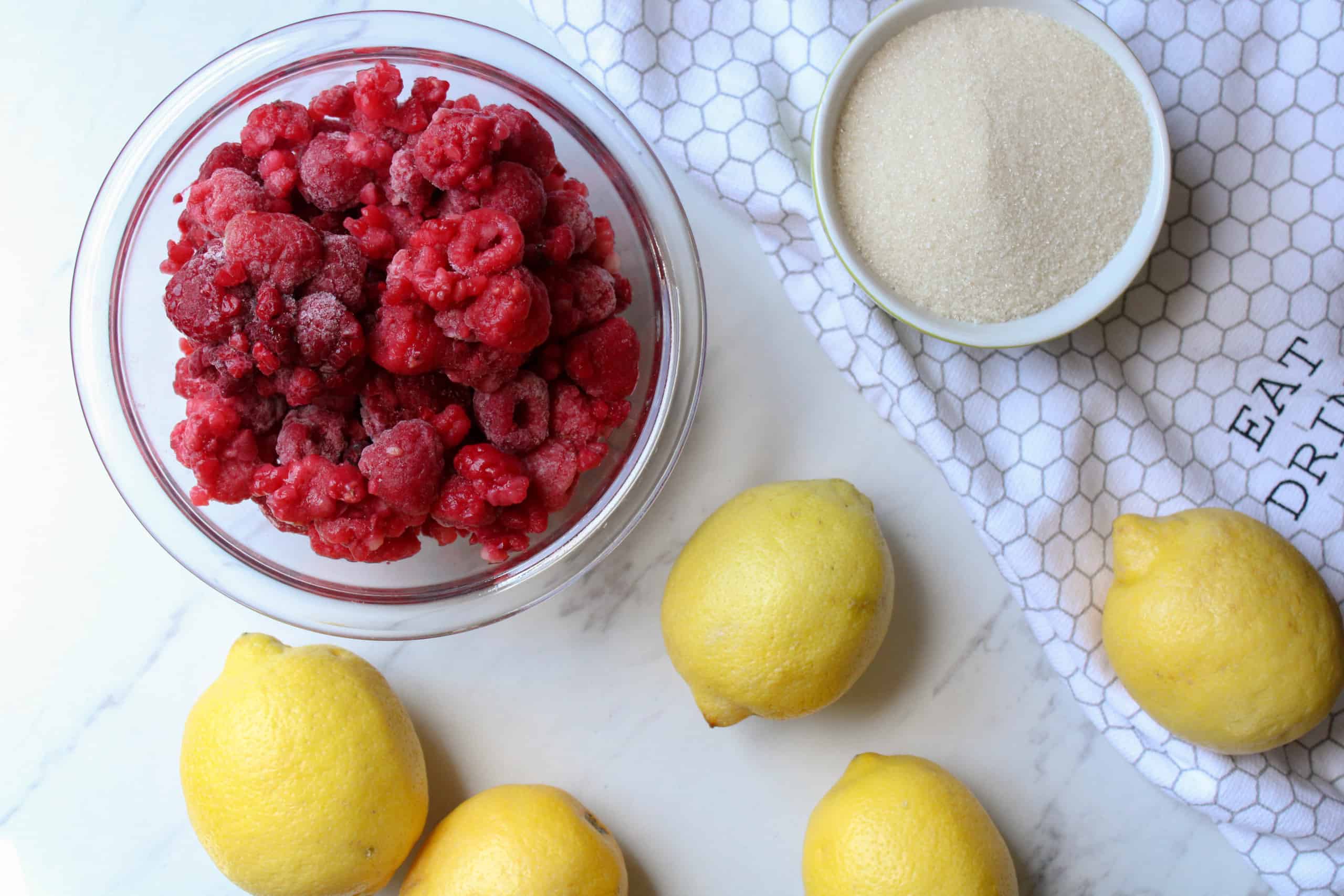 Fresh raspberries, a lemon, and a bowl of sugar arranged on a clean kitchen counter, ready for jam making