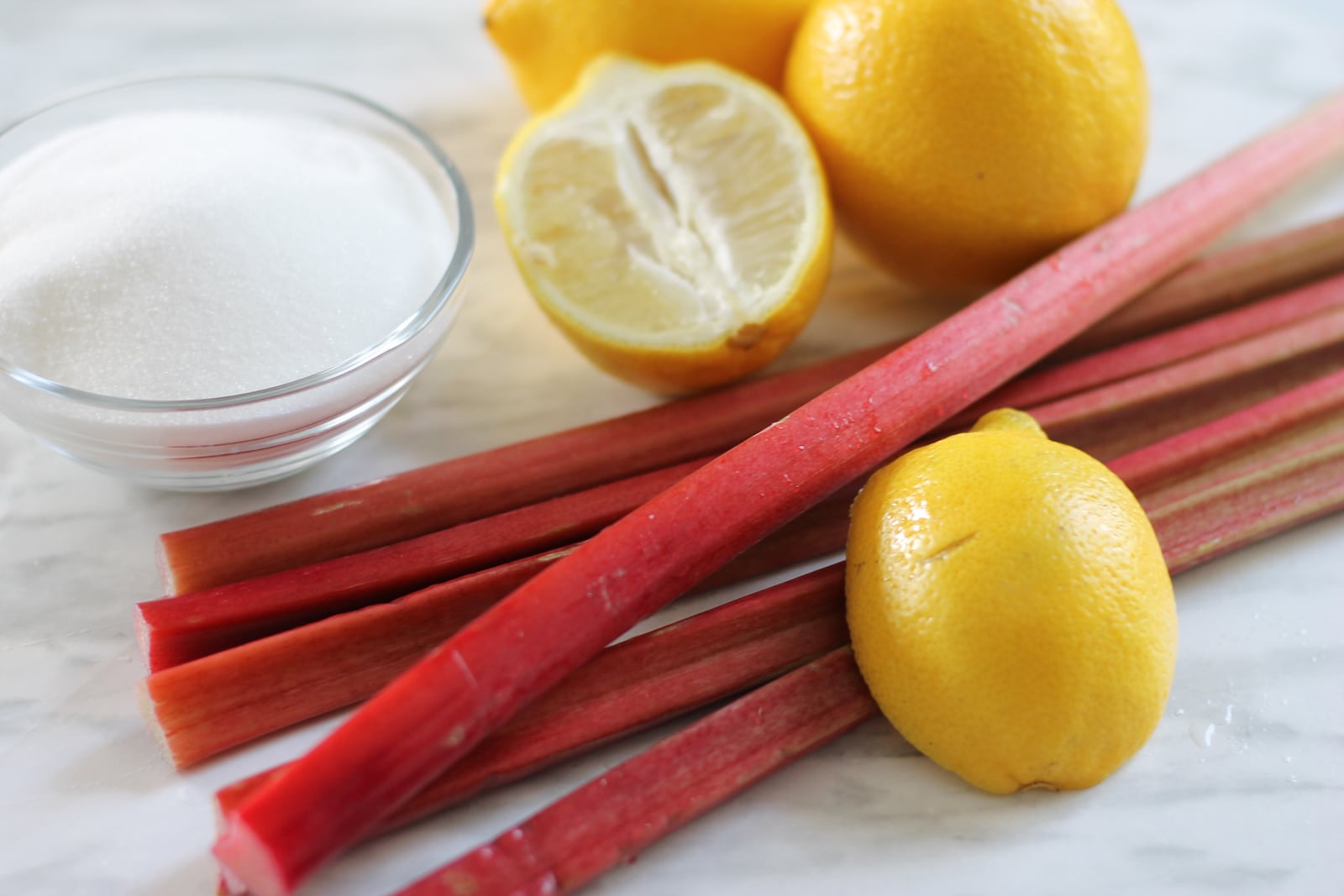 Fresh rhubarb stalks with a small bowl of sugar and a couple of lemons arranged on a wooden cutting board on a kitchen counter, natural window light