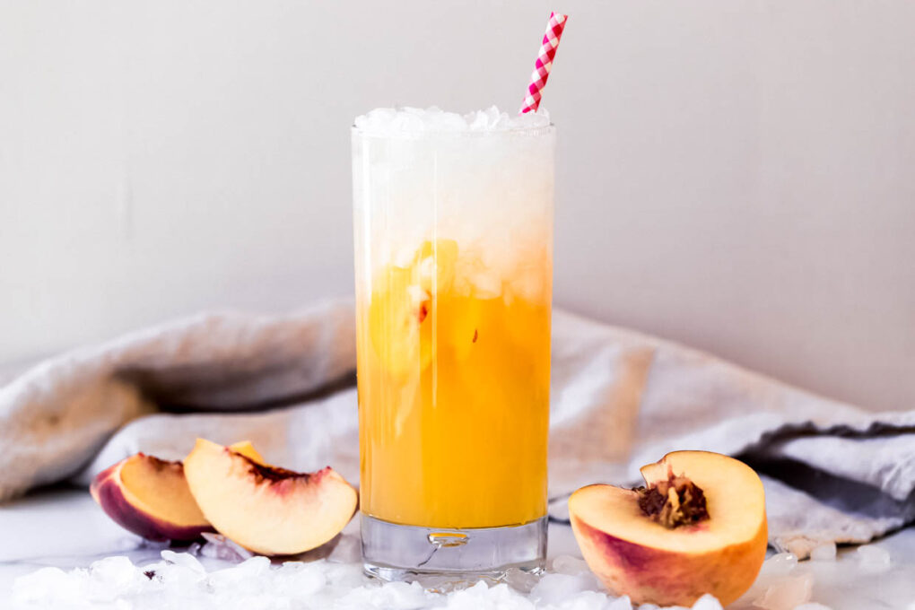 Fresh ripe peaches, a bottle of Prosecco, sugar, and a lemon arranged on a wooden cutting board on a bright kitchen counter