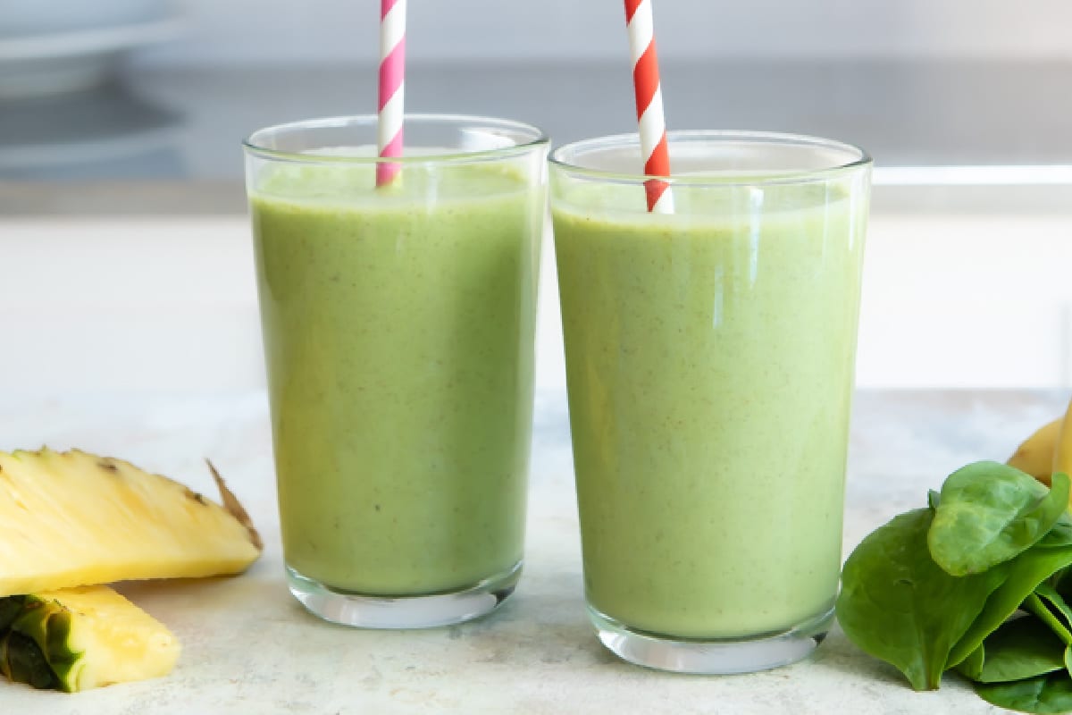 Fresh spinach, a peeled banana, pineapple chunks, Greek yogurt, lemon, and a measuring cup of milk arranged on a kitchen counter