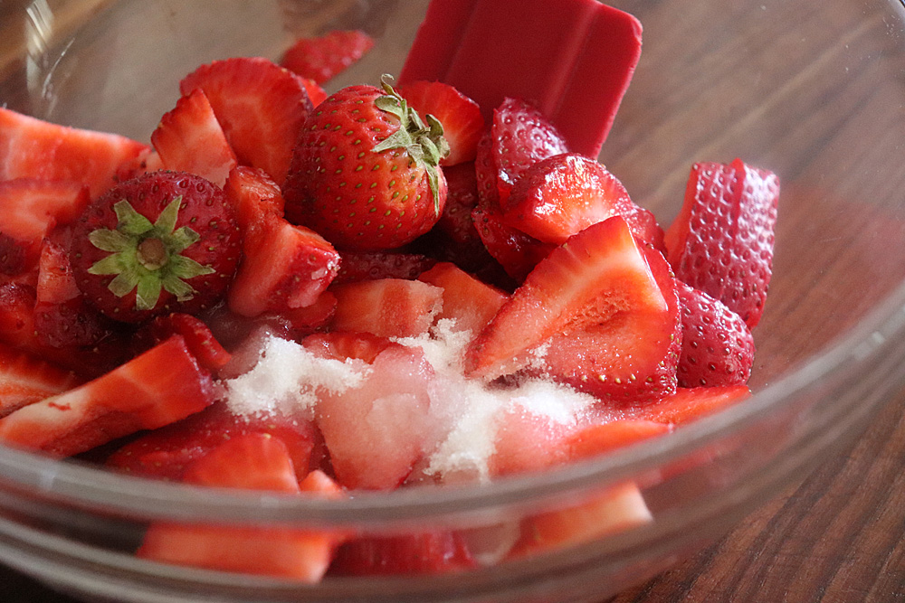 Fresh strawberries, a lemon, granulated sugar, and small bowls of pectin and butter arranged on a tidy kitchen counter in natural light