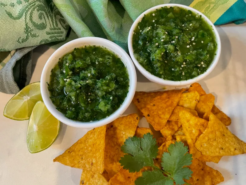 Fresh tomatillos, cilantro, jalapeños, onion, garlic, and limes arranged on a clean kitchen counter with a small bowl of salt