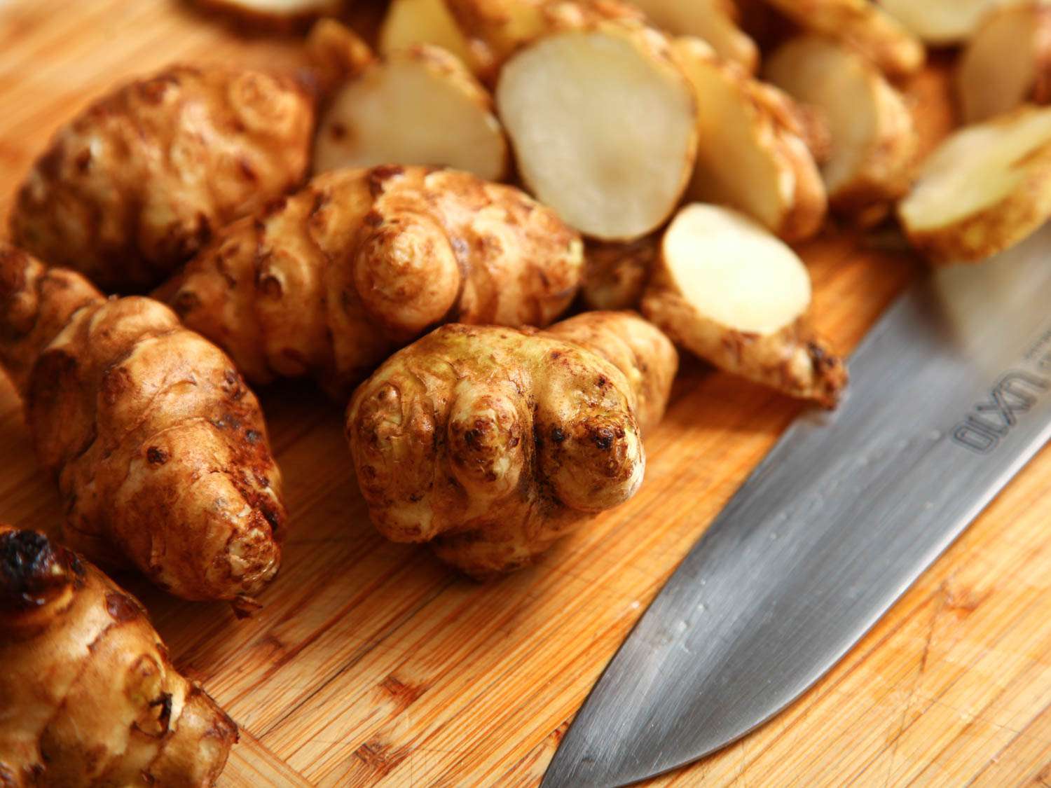 Fresh unpeeled sunchokes on a wooden cutting board next to a chef's knife, with a lemon and a small bowl of kosher salt