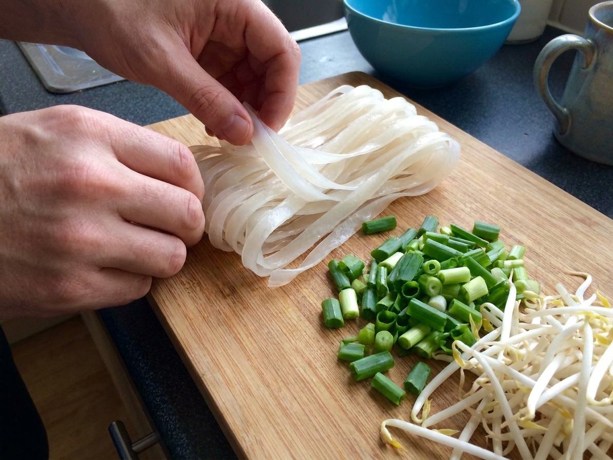 Fresh wide rice noodles being gently separated on a cutting board next to scallions and bean sprouts
